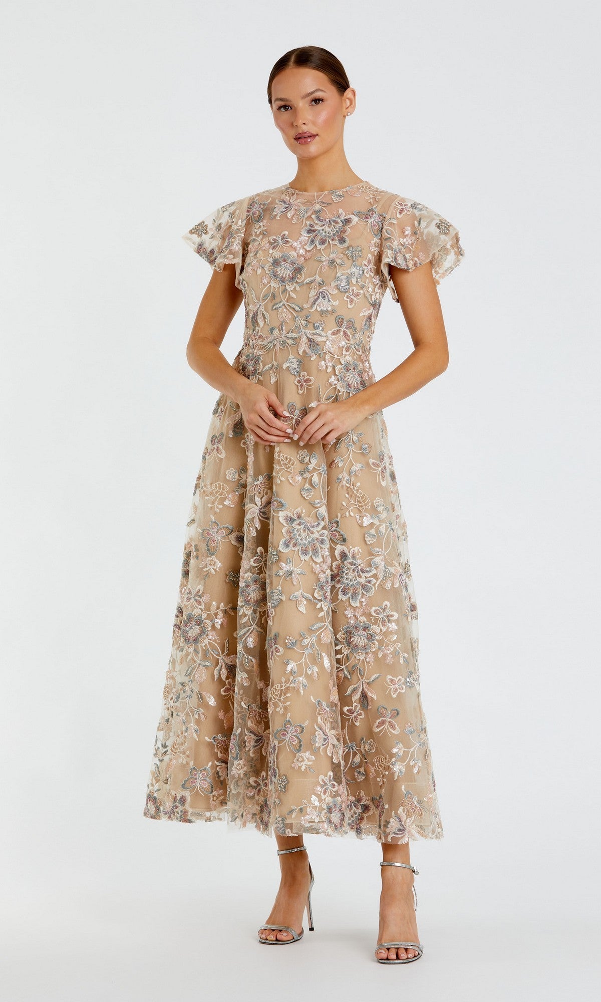 A woman wearing the Mac Duggal 68920 Tea-Length Party Dress in nude, featuring floral embroidery and short flutter sleeves, paired with silver high-heeled sandals, stands against a plain white background.